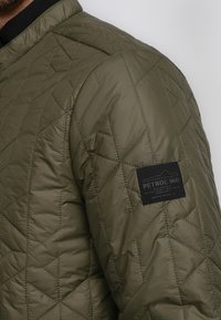 Gepolsterte olivgrüne Jacke mit strukturierter Oberfläche, die ein schwarzes Etikett am Ärmel und gerippte Kragenverzierungen aufweist.