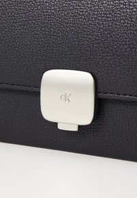 Geantă din piele texturată neagră, având un închizător metallic argintiu cu logo gravat. Finisaj neted, cu formă rectangulară și margini curate.