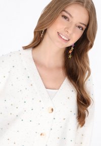 Fehér kardigán színes díszítésekkel, két gombbal és bordázott gallérral. A modellen hosszú haj van és csillag alakú fülbevalót visel.