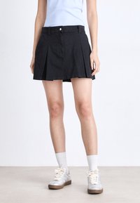 Svart plissé skjørt med en tettsittende midje, laget av bomullsblendstoff. Brukt med grå sneakers og hvite sokker, med et enkelt design.