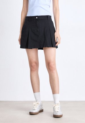 Svart plissé skjørt med en tettsittende midje, laget av bomullsblendstoff. Brukt med grå sneakers og hvite sokker, med et enkelt design.