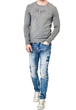 Homme portant un sweat-shirt gris Guess, un jean bleu délavé avec des patches, et des baskets blanches, marchant avec une main dans la poche.