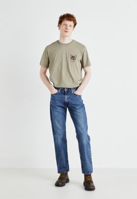 Camiseta de algodón verde oliva con un gráfico de un zorro en el lado izquierdo del pecho, combinada con jeans de pierna recta azul claro y botas marrones con cordones.