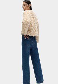 Beige Jacke aus Kunstfell mit cropped Design, kombiniert mit hoch taillierten blauen Jeans aus Denim mit Gesäßtaschen und geradem Bein.