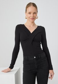 Fekete, hosszú ujjú body V-nyakkivágással és csavart elülső részlettel, magas derekú fekete farmernadrággal, amelyen két hátsó zseb található.