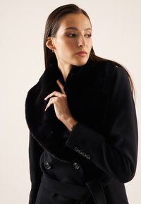 Femme aux longs cheveux bruns portant un manteau noir à double boutonnage avec un large col en fourrure, regardant sur le côté avec la main près du col.