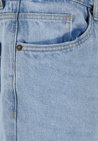 Ljusblå denimjeans med en synlig framficka och kontrasterande gula sömmar. Materialet har en mjuk textur och en lätt urtvättad mönster.