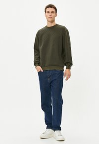 Olijfgroene geribbelde sweatshirt, ronde halslijn, losse pasvorm, gecombineerd met blauwe spijkerbroek en witte sneakers. Eenvoudig, casual ontwerp.