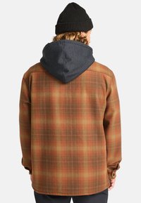 Chaqueta de cuadros en tonos marrón y naranja con una capucha gris. Presenta un corte recto y puños abotonados. Tejido texturizado.