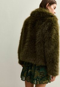 Kobieta w ciemnozielonym sztucznym futrze, nosząca wzorzystą zielono-czarną spódnicę, stoi przodem z głową odwróconą na bok.