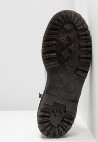 Semelle de botte noire avec des rainures profondes et des motifs en forme de croix, positionnée verticalement contre un mur d'angle blanc sur une surface plane.