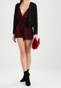 Combinaison bordeaux avec un décolleté en V profond et une taille nouée, associée à une veste courte noire et une pochette en fausse fourrure rouge. Bottines noires.
