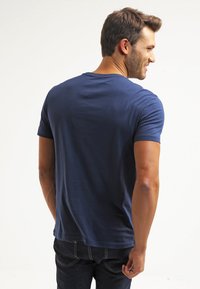 Navy blue cotton t-shirt with short sleeves, crew neck, and a smooth texture, worn with dark denim jeans. Back view highlights fit and style.
