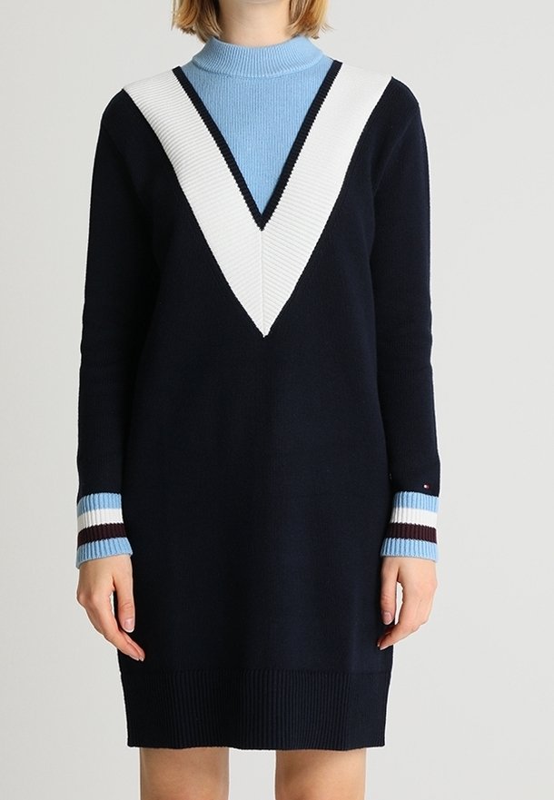 Personne portant une robe-pull bleu marine avec une bande en forme de V blanche et noire sur la poitrine et des poignets rayés bleu clair, blanc et bordeaux.