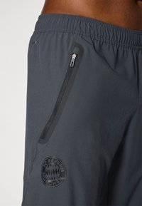 Shorts de sport en matériau gris foncé avec une taille élastique, une poche zippée sur le côté et un logo FC Bayern Munich noir en bas.