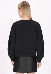 Fekete, oversized pulóver, ejtett vállakkal és bordázott mandzsettákkal, egy fekete bőr mini szoknyával párosítva, hátulról ábrázolva.