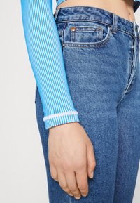 Randig blå långärmad topp med vita manschetter, tillsammans med högmidjade blå denim jeans. Närbild av armen och midjeområdet.