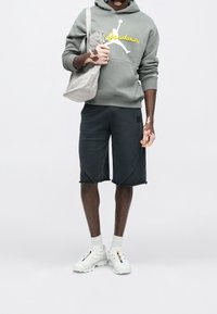 Personne atteinte de vitiligo porte un sweat à capuche gris, un short noir, des baskets blanches et porte un sac fourre-tout gris clair sur un fond uni.