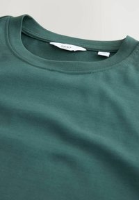 Camiseta de algodón verde oscuro con cuello redondo y etiqueta de marca visible cosida en el interior del cuello, colocada plana sobre una superficie clara.