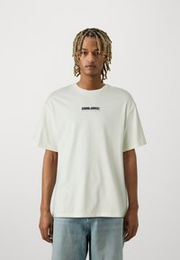 White oversized t-shirt made of cotton, featuring a centered black graphic that reads "SHINJUKU" on the chest. Short sleeves, relaxed fit.