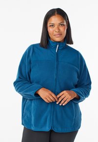 Kvinna med rakt svart hår som bär en blå dragkedjeskjorta av fleece, stående mot en helt vit bakgrund, med händerna nära dragkedjan.