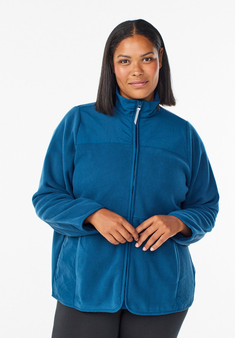 Kvinna med rakt svart hår som bär en blå dragkedjeskjorta av fleece, stående mot en helt vit bakgrund, med händerna nära dragkedjan.