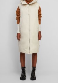 Doudoune longue couleur crème avec fermeture éclair sur le devant et capuche. Portée sur un pull en tricot marron. Bottines noires à lacets avec semelles épaisses.