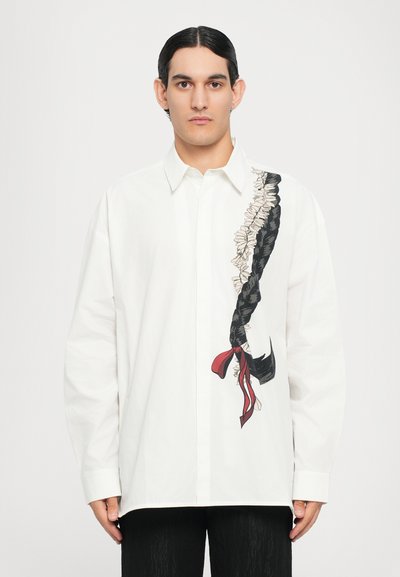 Hvid langærmet skjorte med et grafisk motiv af flettet hår i sort og rød, med blomsteraccenter på siden. Lavet af bomulds- materiale, afslappet pasform.