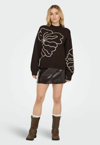 Pull marron avec un grand motif floral blanc, coupe décontractée, manches surdimensionnées, associé à une jupe en cuir noire et des bottes marron montant jusqu'aux genoux.