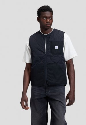 Junger Mann trägt eine schwarze gesteppte Zip-Weste über einem weißen Kurzarmshirt und dunklen Jeans, steht vor einem einfarbigen hellen Hintergrund.