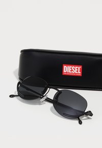 Ochelari de soare ovali negri cu lentile închise și rame subțiri negre, având pusuri de nas clare. Însoțiți de un etui negru cu un logo roșu Diesel.