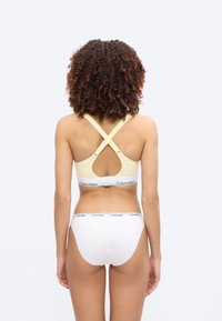 Γυναίκα με σγουρά μαλλιά, φανερή από πίσω, φοράει μπεζ σουτιέν racerback και λευκά εσώρουχα Calvin Klein σε απλό ανοιχτό φόντο.