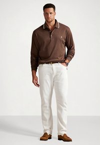 Καφέ μακρυμάνικο πόλο με λευκό γιακά, σε συνδυασμό με λευκό παντελόνι και καφέ μπλεϊζερ σουέντ παπούτσια. Ορατό ρολόι στον καρπό. Ουδέτερο φόντο.