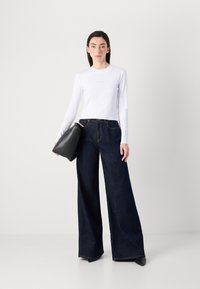 Una giovane donna sta in piedi, indossando un top bianco a maniche lunghe aderente e jeans scuri a gamba larga, mentre tiene una borsa nera contro uno sfondo neutro.