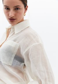 Lekka, przezroczysta koszula zapinana na guziki w kolorze kremowym, z wyraźnym kołnierzykiem, dwiema kieszeniami na piersi oraz luźnym krojem z delikatną teksturą.