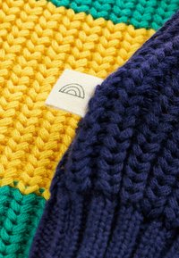 Coperta lavorata a maglia con sezioni vivaci in giallo, verde e blu. Un bordo blu navy con un'etichetta di stoffa che mostra un logo arcobaleno è visibile.