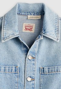 Ανοιχτό μπλε τζιν μπουφάν με μυτερό γιακά, δύο τσέπες στο στήθος, ασημένια κουμπιά και κόκκινη ετικέτα εμπορικού σήματος μέσα στον λαιμό.