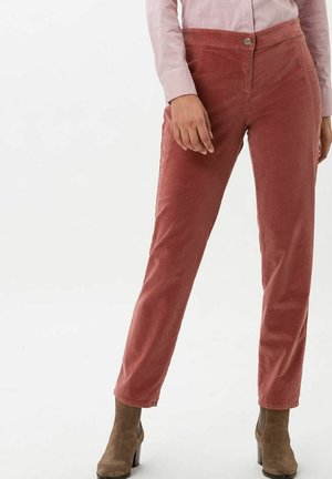 Frau trägt staubrosafarbene Cordhosen, ein hellrosa gestreiftes Hemd und braune Wildleder-Stiefeletten mit Absatz, steht vor einem schlichten Hintergrund.