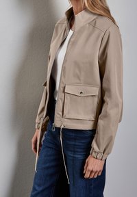 Veste légère beige avec fermeture éclair, col montant, poignets élastiques et poche frontale, associée à un jean bleu foncé.