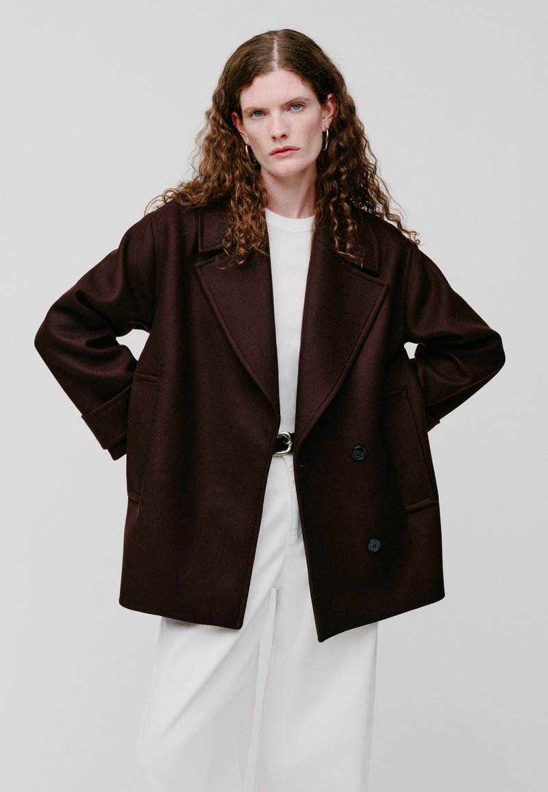 Cappotto oversize marrone scuro con ampi revers, doppi bottoni e tasche laterali, indossato sopra una maglietta bianca e abbinato a pantaloni bianchi.