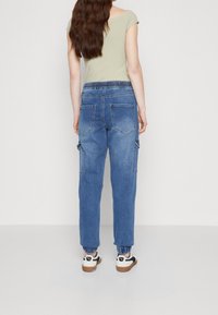 Kvinna med långt brunt hår som bär en grön kortärmad topp, blå denim cargobyxor med elastisk midja och vita sneakers med gummisula, stående vänd bort.