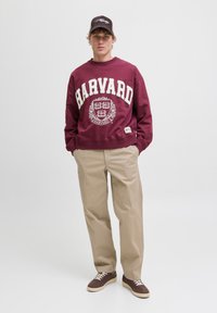 Sudadera burdeos con el texto "HARVARD" en blanco llamativo, pantalones beiges, zapatillas marrones y una gorra oscura. Diseño sencillo, ajuste relajado, material de algodón.