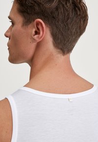 Witte tanktop met een ronde halslijn en zichtbare stiksels. Gladde textuur met een klein label aan de achterkant. Details in lichtgrijs.