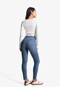 Haut à manches longues blanc côtelé, jean skinny bleu taille haute et baskets blanches avec des accents marron. Vue arrière, textures lisses, design ajusté.