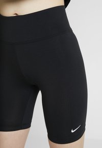 Närbild av midjan och övre låren iförda svarta Nike cykelshorts med hög midja och vit Nike swoosh-logotyp på vänster ben.