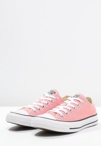 Para niskich różowych trampków z płótna, z białymi gumowymi podeszwami i sznurówkami, z czarnymi paskami i etykietami na języku z logo.