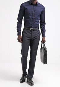 Marinblå skjorta, slim-fit grå byxor, svart läderbälte och skor. Man håller en svart läderväska, står mot en enfärgad bakgrund.