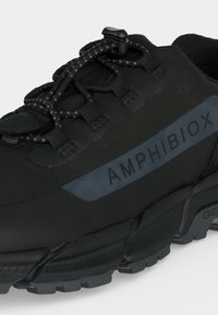 Chaussure de plein air noire avec semelle à épaisse bande de roulement, lacets élastiques avec bouton-pression, et "AMPHIBIOX" imprimé sur le panneau latéral.