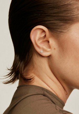 Pendientes de plata con un diseño floral y una superficie texturizada, colocados en un primer plano de una oreja, con cabello corto de color oscuro visible.