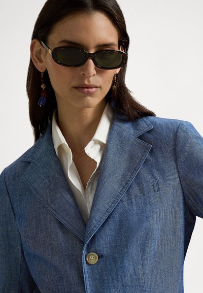 Rifľové sako zo svetlomodrej látky s klopám a jedným gombíkom. Biela košeľa pod ním. Hnedé slnečné okuliare s motívom korytnačky a farebné náušnice.